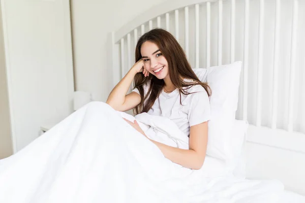 Attraktive junge Frau lächelt und blickt in die Kamera, während sie unter der Decke im Bett sitzt — Stockfoto