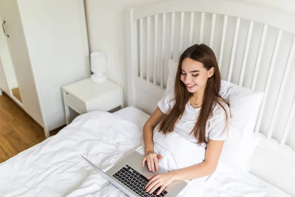 Mujer morena joven feliz usando el ordenador portátil mientras está sentado en la cama por la mañana — Foto de Stock