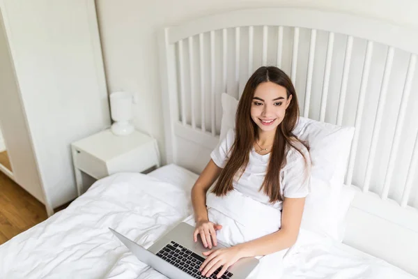 Mujer morena joven feliz usando el ordenador portátil mientras está sentado en la cama por la mañana — Foto de Stock