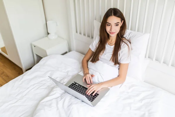 Mujer joven usando computadora portátil en el dormitorio. Modelo femenino en la cama trabajando en el ordenador portátil . — Foto de Stock