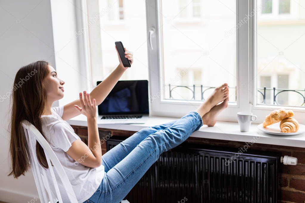 Beautiful young woman sitting on chair with legs on windowsill, laughing and taking selfie on the phone happily in the morning breakfast time