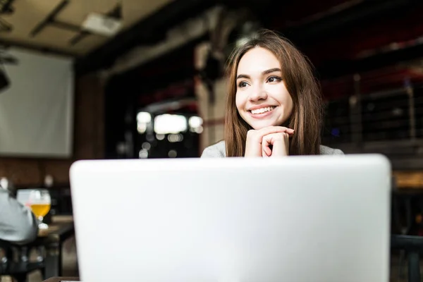 Vrij vrouwelijke student met schattige glimlach keyboarding iets op net-boek terwijl u ontspant na lezingen in universiteit. Mooie gelukkige vrouw die werkt op laptopcomputer tijdens de koffiepauze in café-bar — Stockfoto