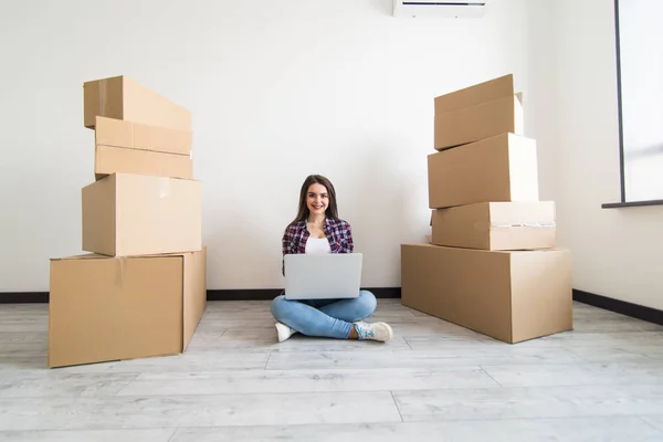 Attraktiv ung kvinna är rörligt sittande bland kartonger, använder en bärbar dator i nya lägenheten — Stockfoto