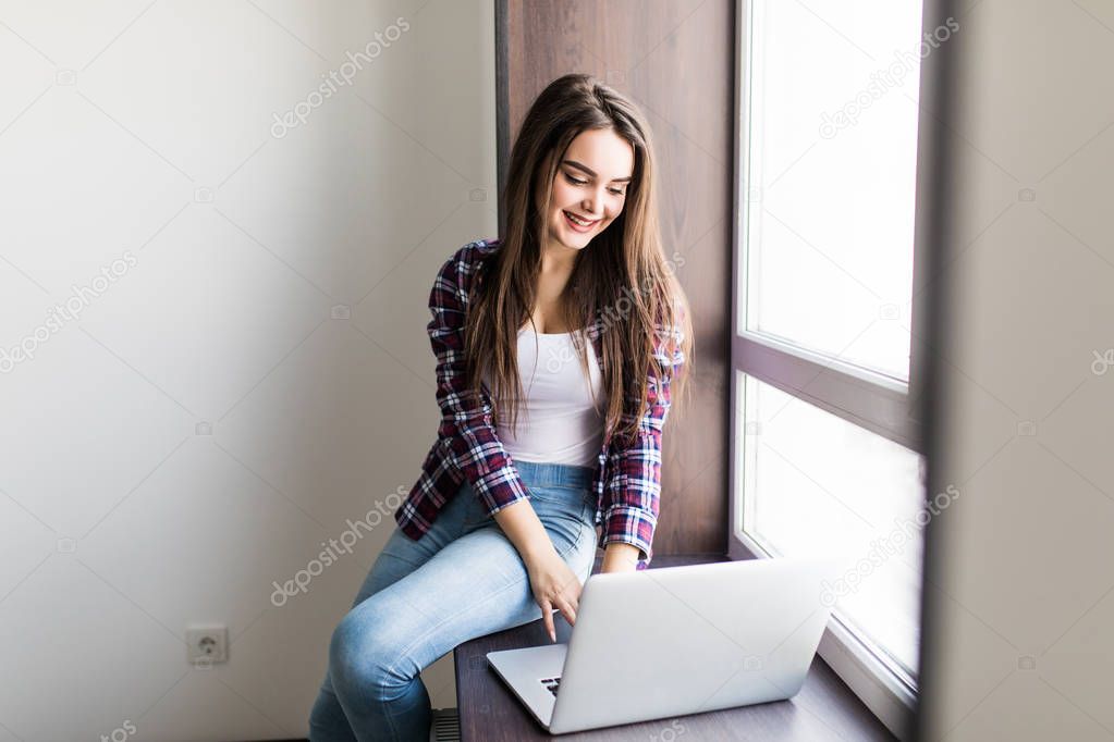 Portrait of a stylish and young beautiful woman sitting with a laptop on her lap on the windowsill, leaning against the window
