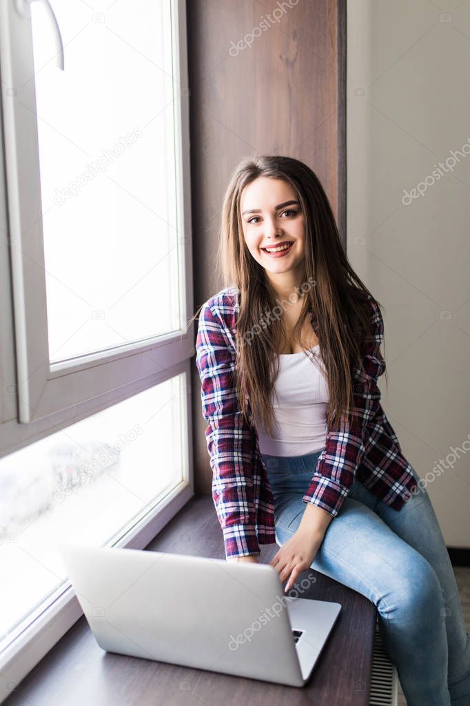 Portrait of a stylish and young beautiful woman sitting with a laptop on her lap on the windowsill, leaning against the window