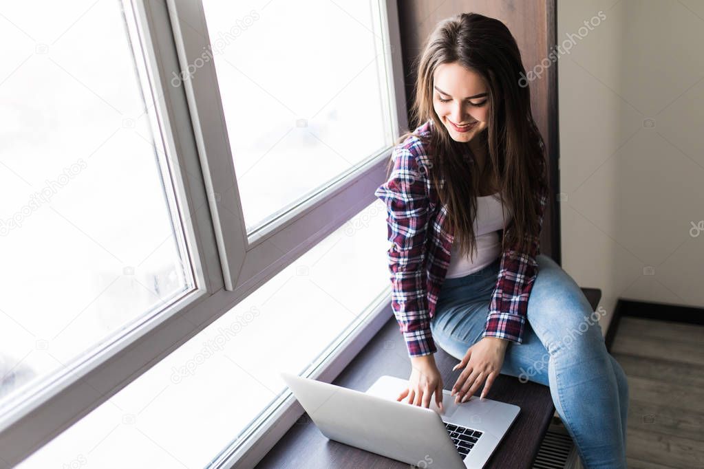 Portrait of a stylish and young beautiful woman sitting with a laptop on her lap on the windowsill, leaning against the window