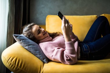 Evdeki oturma odasındaki kanepede uzanmış akıllı bir telefonla rahatlamış bir kızın fotoğrafını çek.