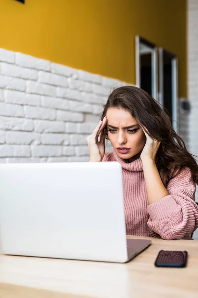 Retrato de uma mulher atraente à mesa com laptop com as mãos em seus templos de dor de cabeça . — Fotografia de Stock