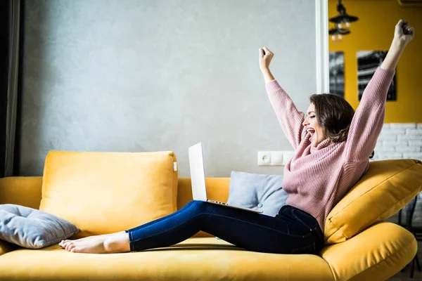 Young woman raising her hands up on the couch with laptop at home — Stock Photo, Image