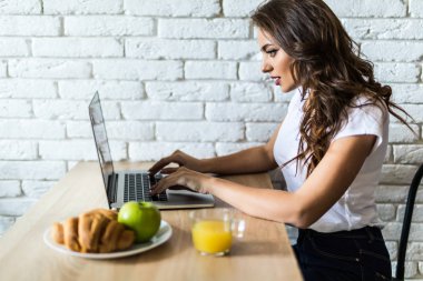 Dizüstü bilgisayar kullanan ve sabah mutfakta meyve yeme genç neşeli kadın