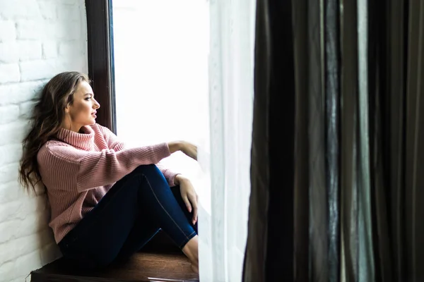 Smiling pretty woman in sweater sitting on a windowsill at home — Stock Photo, Image