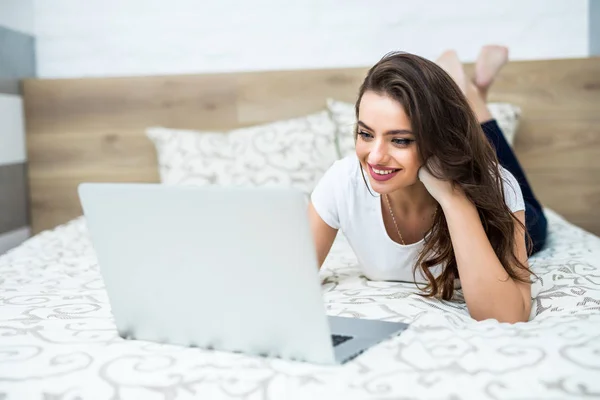 Uśmiechnięta kobieta z jej nogi lekko podniesiona przed jej laptop leżąc na łóżku. — Zdjęcie stockowe
