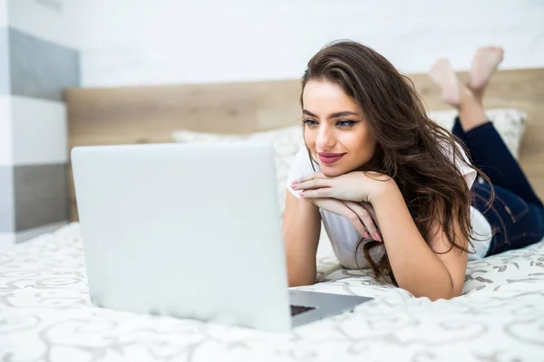 Mujer sonriente con las piernas levantadas levemente delante de su computadora portátil acostada en la cama . — Foto de Stock
