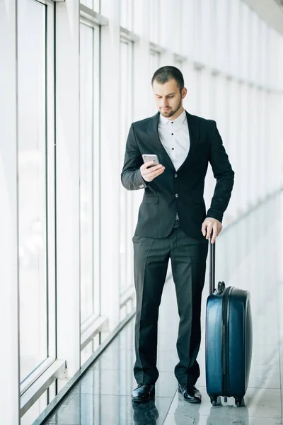 Glücklicher junger Geschäftsmann, der am Flughafen auf sein Handy schaut. gutaussehender Geschäftsleiter, der auf dem Smartphone SMS schreibt, während er im Flughafenterminal spaziert. — Stockfoto