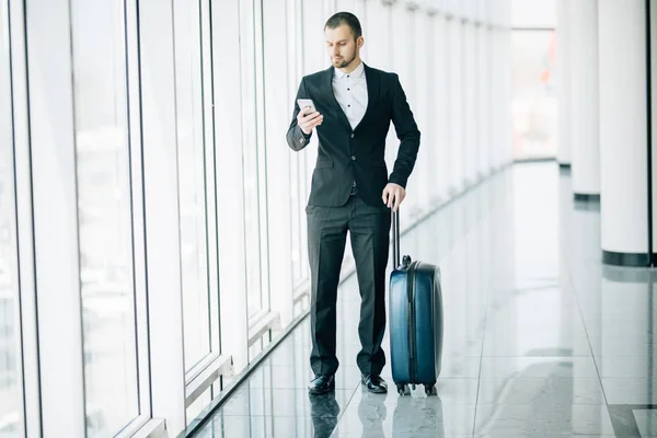 Elegante empresário verificando e-mail no telefone celular enquanto caminha com mala dentro do terminal do aeroporto. Empregador experiente do sexo masculino usando telefone celular antes de viajar para o trabalho — Fotografia de Stock