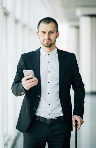 Mladý podnikatel s kufrem a za použití chytrého telefonu čeká na let. Kavkazská podnikatel čekání na terminálu držet mobilní telefon. — Stock fotografie