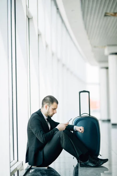 Jovem homem de negócios sentado ao telefone com a mala no aeroporto esperando o voo atrasado — Fotografia de Stock