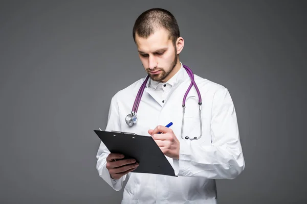 Portræt af skægget smilende læge praktikant holder udklipsholderen med tomt papir. Doc har hvid uniform på på en lysegrå baggrund. - Stock-foto