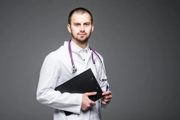 Retrato de un interno médico sonriente barbudo sosteniendo el portapapeles con papel vacío. Doc lleva puestos uniformes blancos sobre un fondo gris claro —  Fotos de Stock