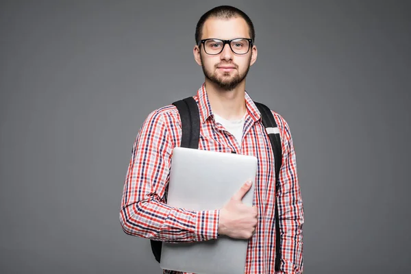 Bonito e jovem estudante universitário masculino levando saco no fundo branco enquanto segurando laptop faculdade isolado no cinza — Fotografia de Stock