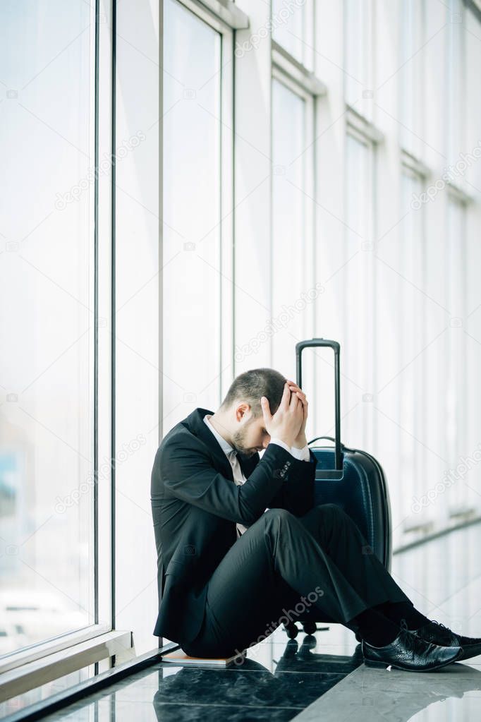 Business man sitting at the terminal airport on the floor with suitcase flight delay, two hands touch at head, headache, waiting traveling. Business trip