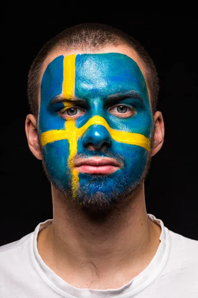 Portrait of handsome man face supporter fan of Sweden national team with painted flag face isolated on black background. Fans emotions.
