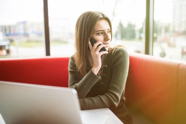 Dizüstü bilgisayar ve telefon kafede kullanan mutlu kadın. Bir kafede otururken ve bilgisayarda çalışan genç güzel kız