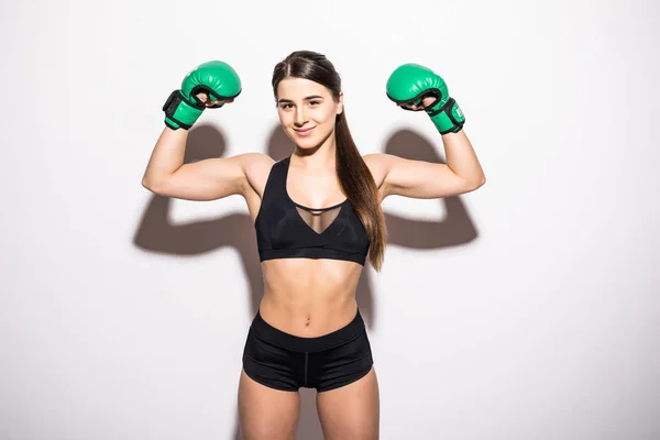 Lachende brunette fitness vrouw in bokshandschoenen weergeven van haar biceps tijdens het kijken naar de camera op witte achtergrond — Stockfoto