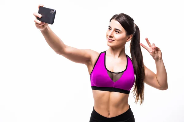 Vackra leende passar idrottskvinna tar en selfie och visar fred gest stående över vit bakgrund — Stockfoto