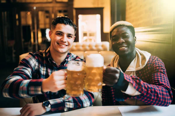 Dos mejores amigos o compañeros de universidad tomando cerveza en el pub. Afro hombre americano hablando con su amigo caucásico en el bar — Foto de Stock
