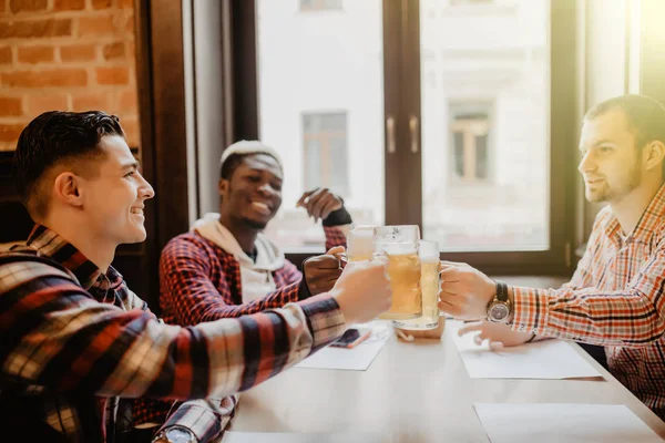 Guapos hombres multiétnicos están tintineando taza de cerveza hablando sonriendo mientras descansan en el pub — Foto de Stock
