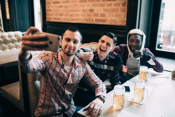 Multiracial vänner grupp i casual kläder leende, tar selfie och dricka öl medan du sitter i puben — Stockfoto