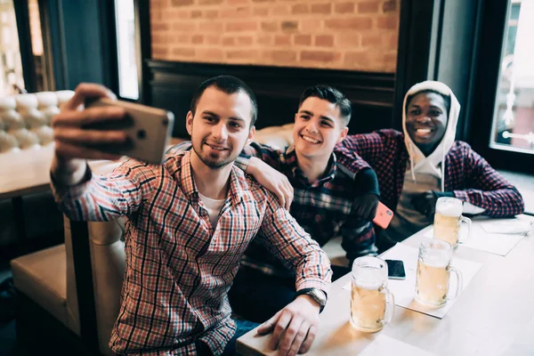Tre unga män i casual kläder leende, ta selfie och dricka öl medan du sitter på pub — Stockfoto