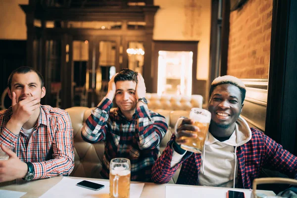 Titta på Tv i baren. Tre lyckliga unga män dricker öl och gestikulerande med olika känslor när man sitter i bar stöd annan klubb eller team — Stockfoto