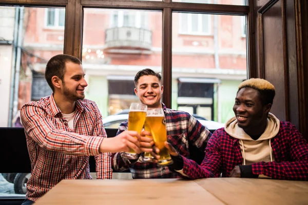 Salud, amigos míos. Tres hombres guapos multirraciales están bebiendo cerveza, celebrando la reunión y sonriendo . — Foto de Stock