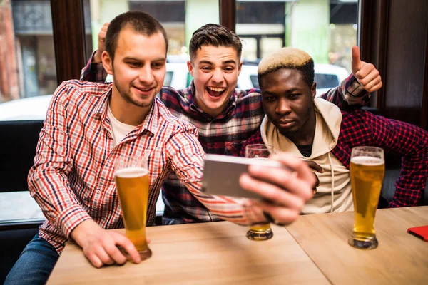 Three multiracial young men in casual clothes are taking selfie and drinking beer while sitting in pub