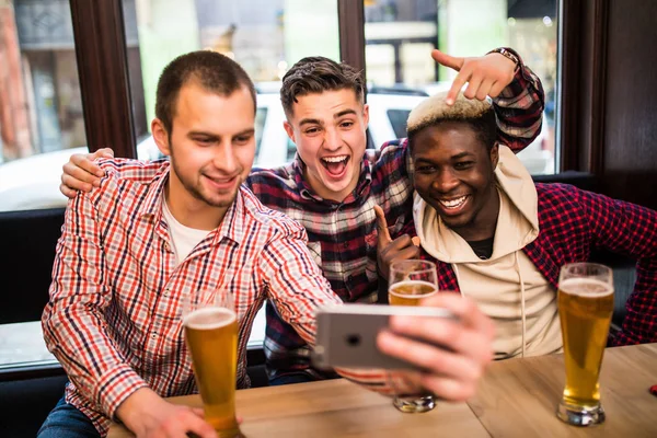 Three multiracial young men in casual clothes are taking selfie and drinking beer while sitting in pub — Stock Photo, Image