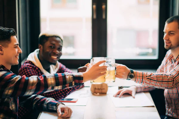 Felices amigos masculinos multirraciales bebiendo cerveza y tintineando vasos en el pub — Foto de Stock