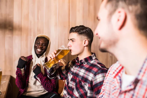 Tres hombres multirraciales con ropa casual sonríen y beben cerveza mientras están sentados en el mostrador del bar en el pub — Foto de Stock