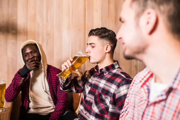 Três homens multirraciais em roupas casuais estão sorrindo e bebendo cerveja enquanto estão sentados no balcão do bar no pub — Fotografia de Stock