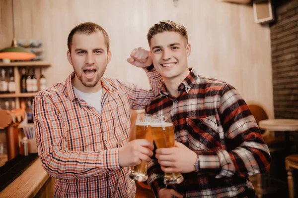 A ver futebol no bar. Dois jovens bonitos de uso casual segurando óculos com cerveja e olhando para longe enquanto estavam no balcão do bar — Fotografia de Stock