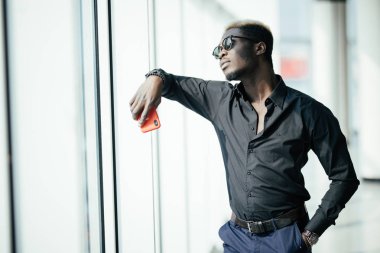 Yakışıklı Afro Amerikalı işadamı rahat kıyafetler ve gözlük panoramik pencerelere karşı cep telefonu tutuyor