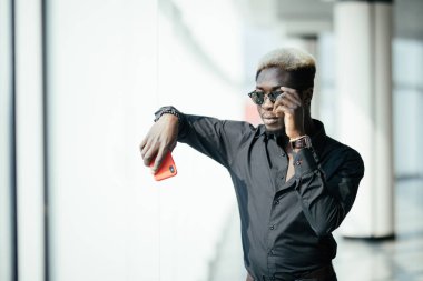 Yakışıklı Afro Amerikalı işadamı rahat kıyafetler ve gözlük panoramik pencerelere karşı cep telefonu tutuyor