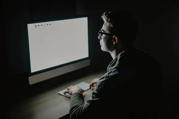 Primer plano del joven sentado y el uso de la computadora de pantalla en blanco en el cuarto oscuro. hacker o programador en cuarto oscuro . — Foto de Stock
