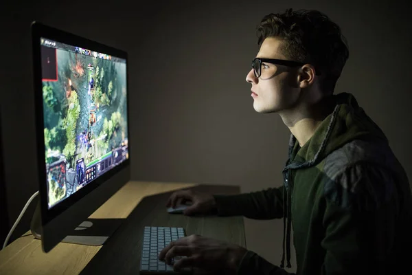 Jogo nocturno. jovem gamer em fones de ouvido e óculos usando o computador para jogar jogo em casa na noite — Fotografia de Stock