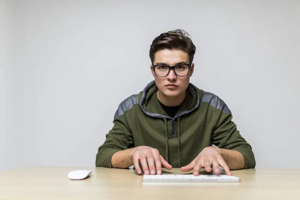 Jeune hacker avec des lunettes vêtu de vêtements décontractés assis à un bureau et travaillant sur un clavier d'ordinateur devant — Photo