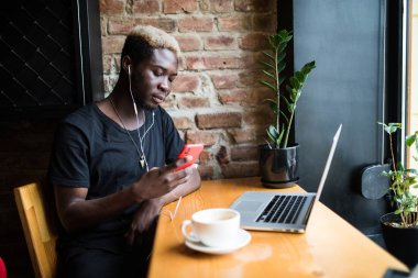 Kafede oturan, müzik dinlemek ve telefon ekranına bakarak yakışıklı afro Amerikalı adam