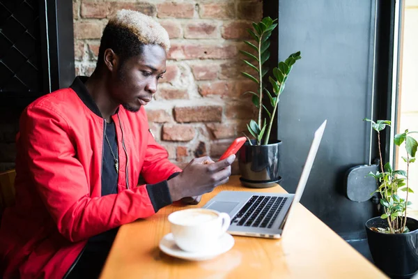 Guapo chico afroamericano sentado en la cafetería con el teléfono en las manos delante de la computadora portátil abierta, el teclado y la búsqueda de Internet, beber café. Joven estudiante de piel oscura preparándose para la clase — Foto de Stock