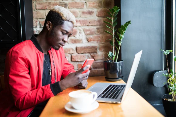 Un joven africano sentado en la cafetería enviando mensajes de texto desde su teléfono móvil. Estudiante vistiendo ropa de moda, tomando café, usando Internet inalámbrico. Concepto de tecnología y comunicación — Foto de Stock