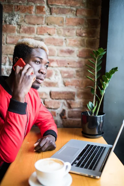 Un joven africano sentado en la cafetería habla del teléfono móvil. Estudiante vistiendo ropa de moda, tomando café, usando Internet inalámbrico. Concepto de tecnología y comunicación — Foto de Stock